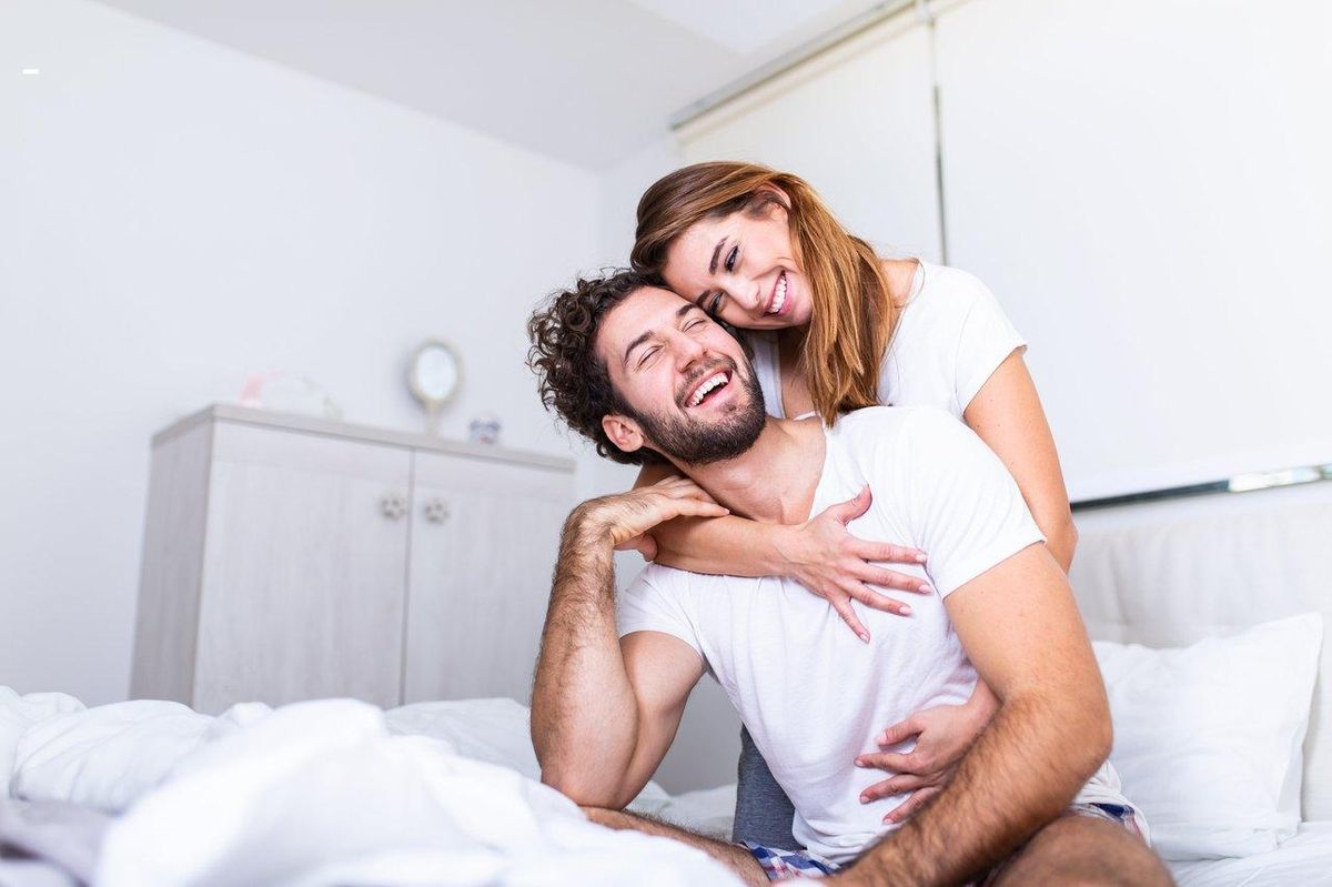 Un couple souriant et complice dans leur chambre, explorant la intimité avec des sextoys pour pimenter leur relation.
